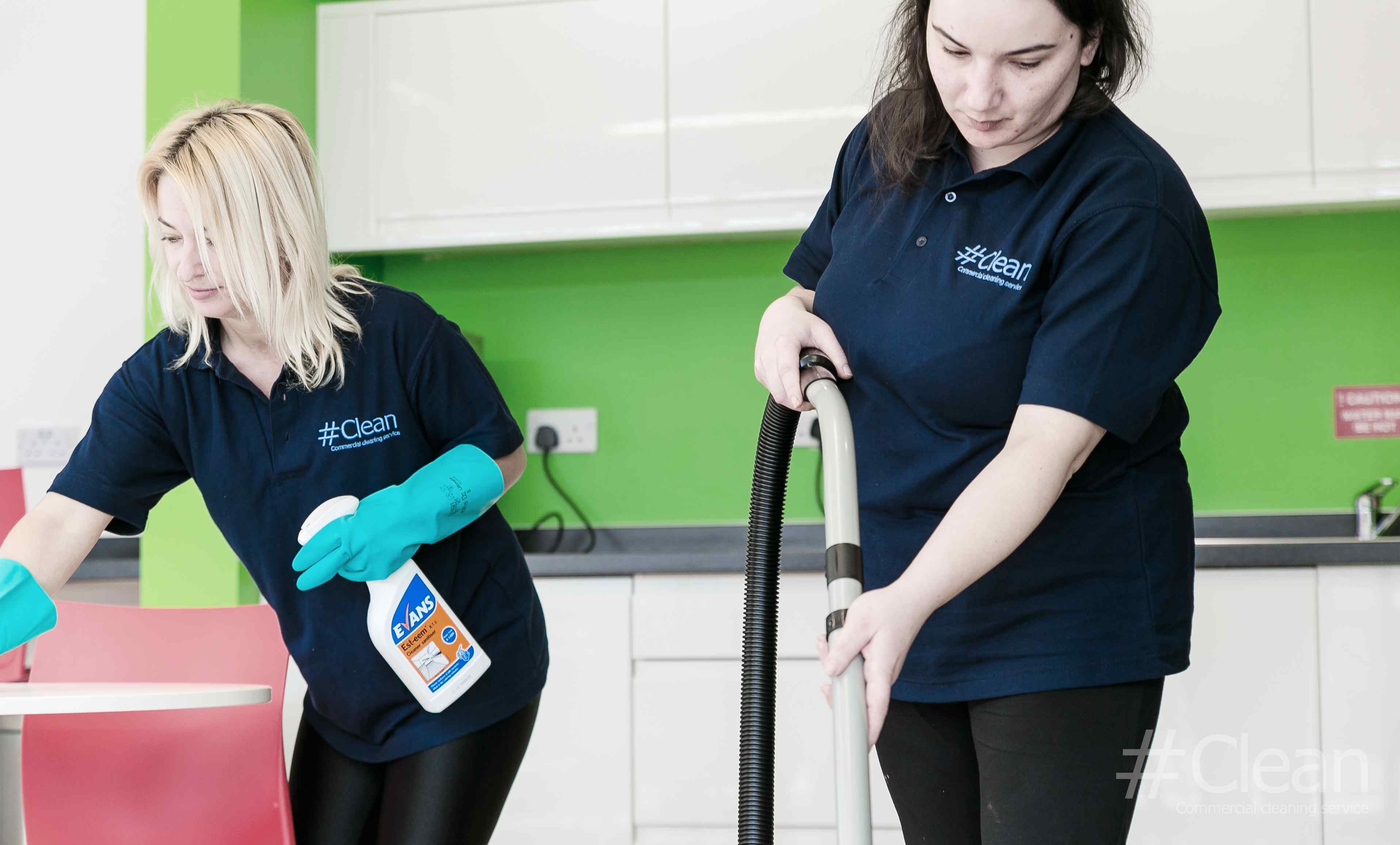 Two Women Cleaning One Is Hoovering And The Other Is Using Cleaning Spray