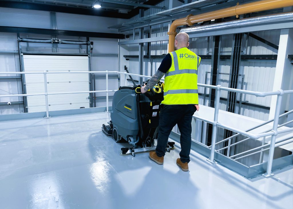 Hashtag Clean team member operating an industrial floor cleaning machine in a modern warehouse or commercial facility while wearing a branded high-visibility vest.