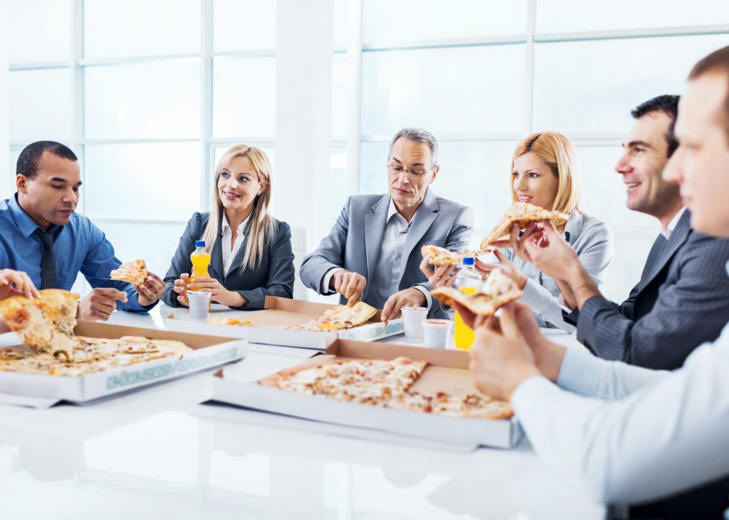 Business professionals sitting around a table in an office, enjoying pizza and drinks during a casual team lunch or meeting.